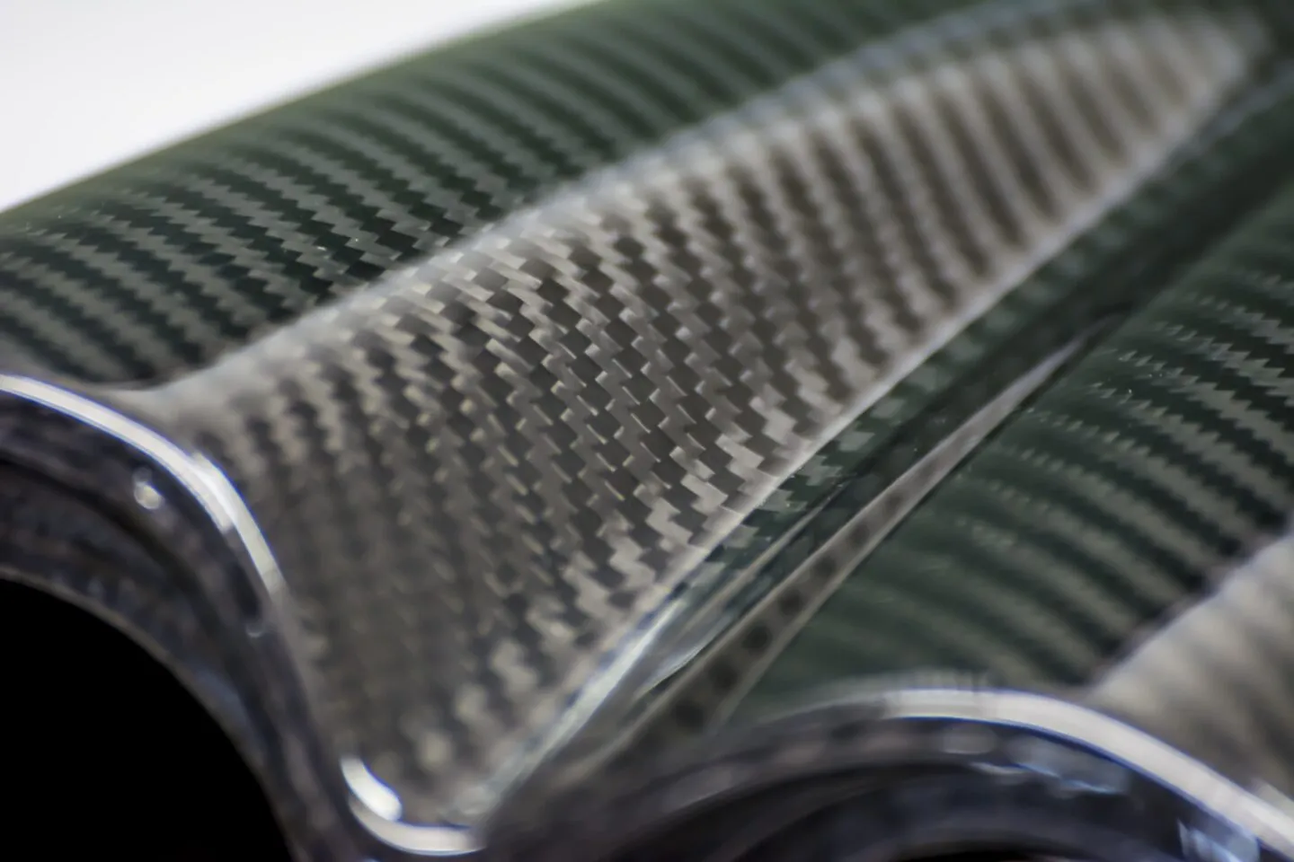 Close-up of a sleek carbon fiber material with a shiny, woven texture. The curved surface reflects light, emphasizing its high-tech, modern appearance.